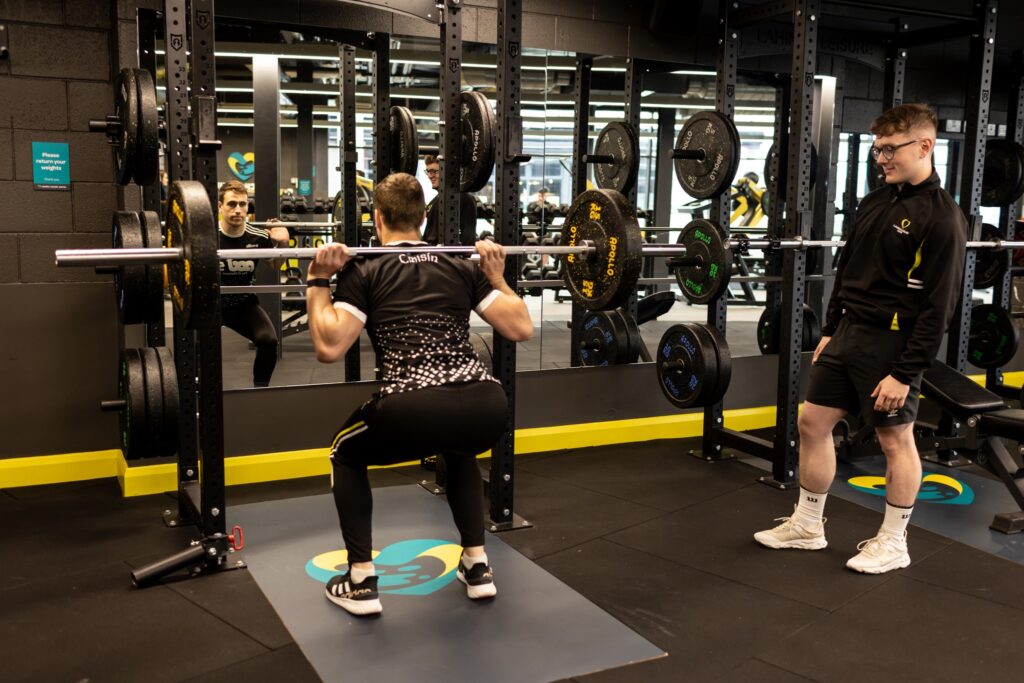 A man is performing a barbell squat in a gym while another man, likely a trainer, watches and supports him. The gym has mirrors and weight plates on the rack. The man squatting is wearing black athletic wear, and the trainer is dressed in a black jacket and shorts.