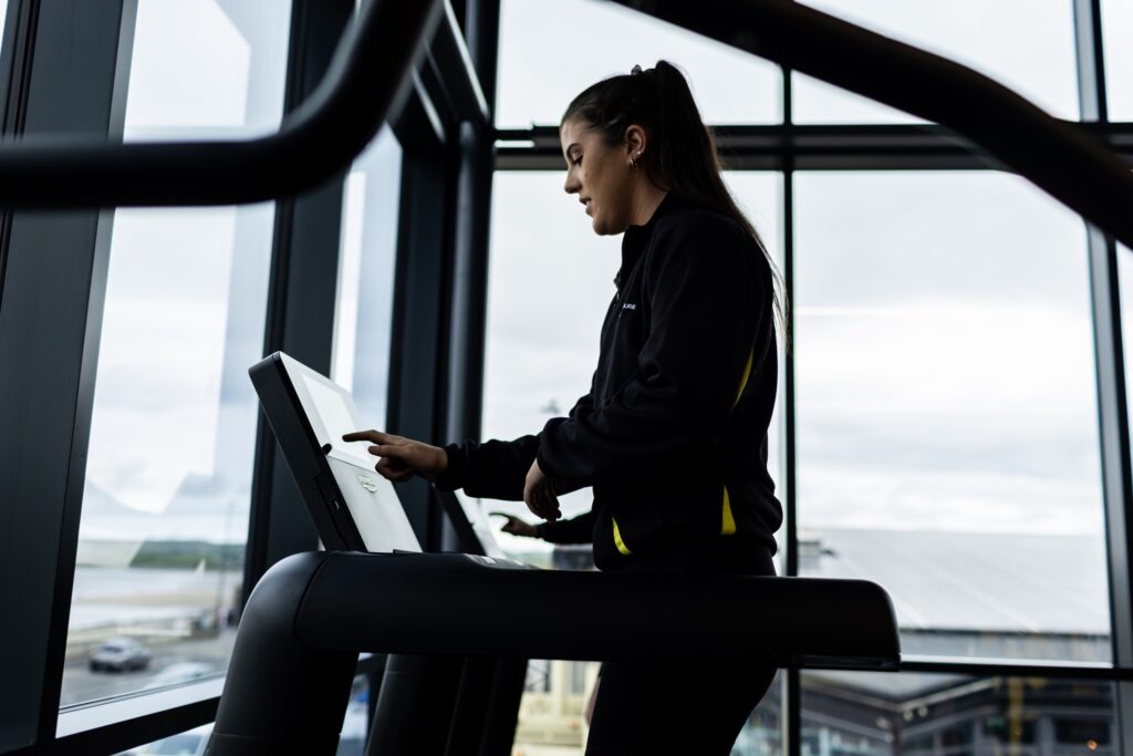 Side view of a woman with a ponytail using the touch screen on a treadmill in a gym with large windows showing an outdoor view.