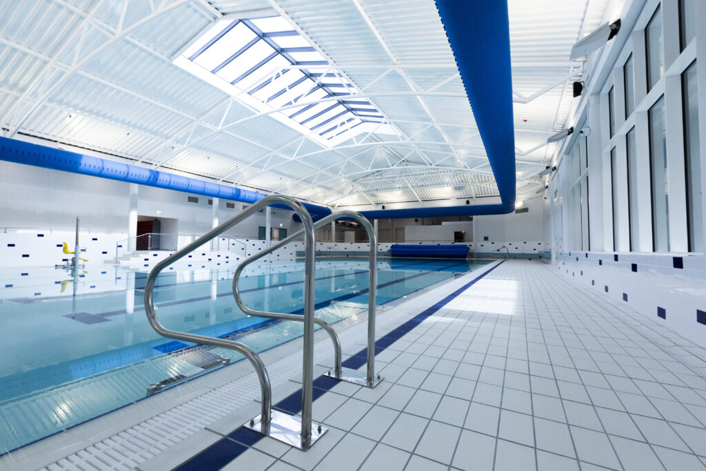 Indoor swimming pool with clear blue water, stainless steel handrails, white tiled floor and walls, bright skylights, and blue ventilation ducts along the ceiling.